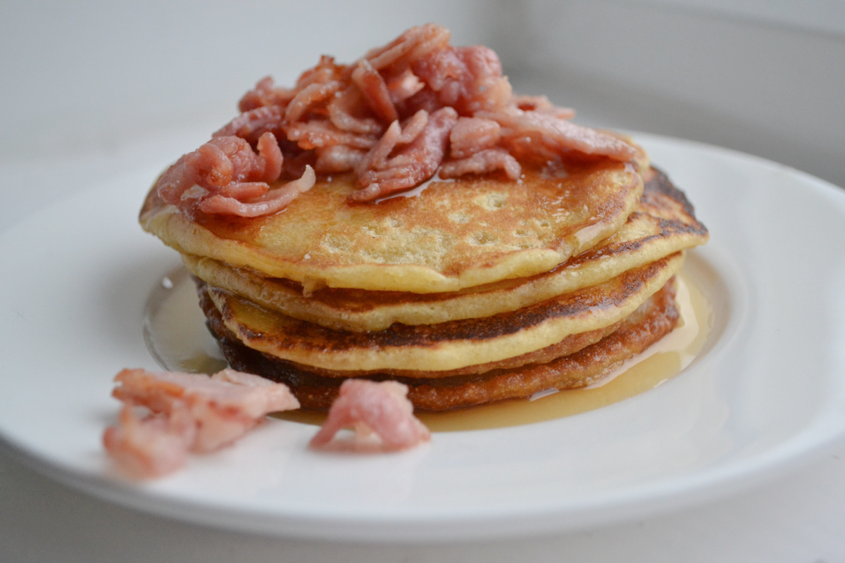 A stack of pancakes in a white bowl with maple syrup and bacon pieces