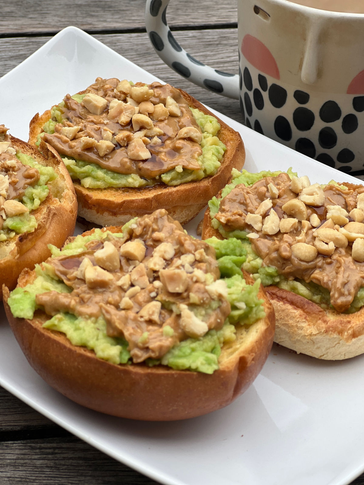4 brioche toasted buns with avocado and peanut butter on top with a black and white mug of tea on a wooden table and a white plate